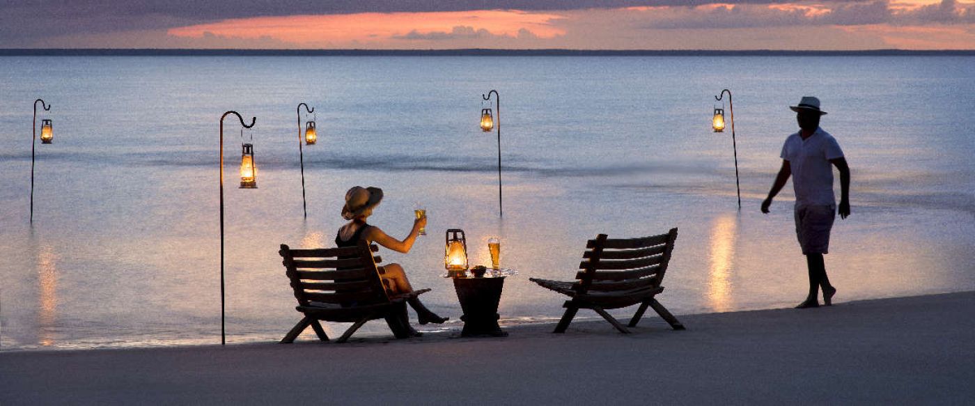 man walking towards woman having lantern lit sun-downers on a beach